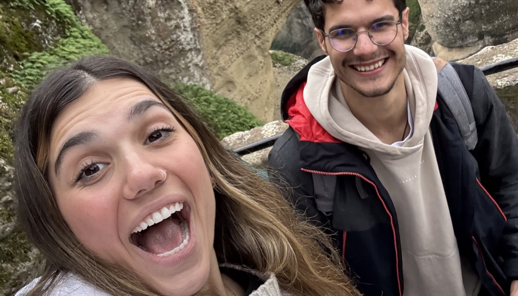 An excited young woman taking a selfie with a smiling man wearing glasses outdoors near rocky cliffs.