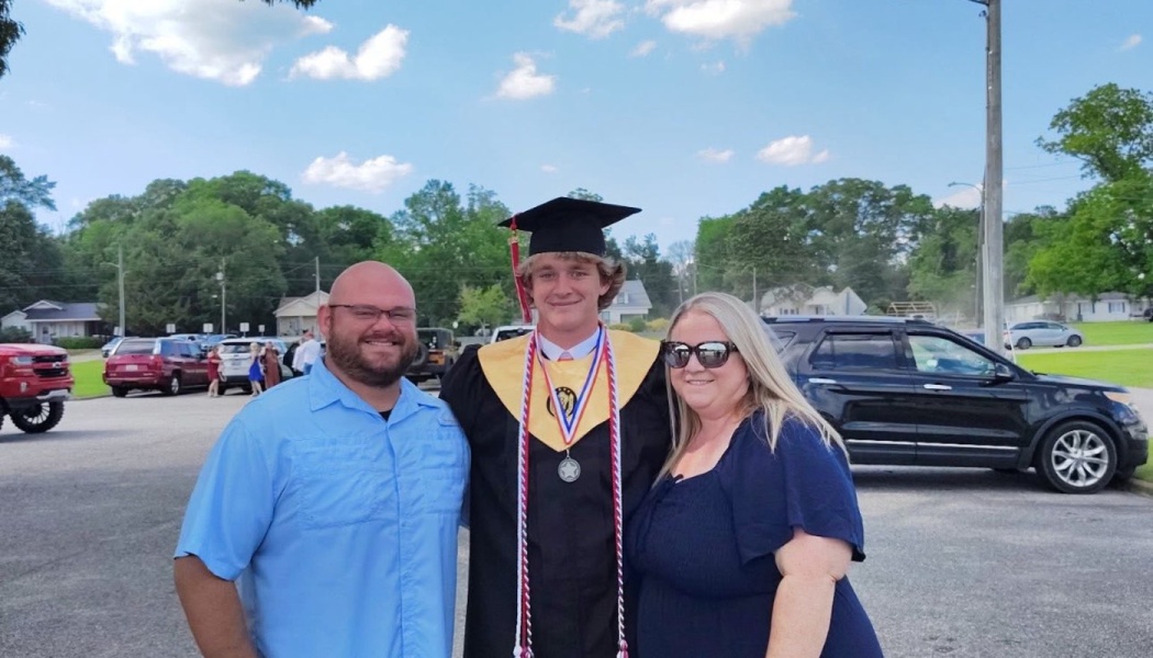 A smiling graduate in a black cap and gown stands outdoors between his parents.