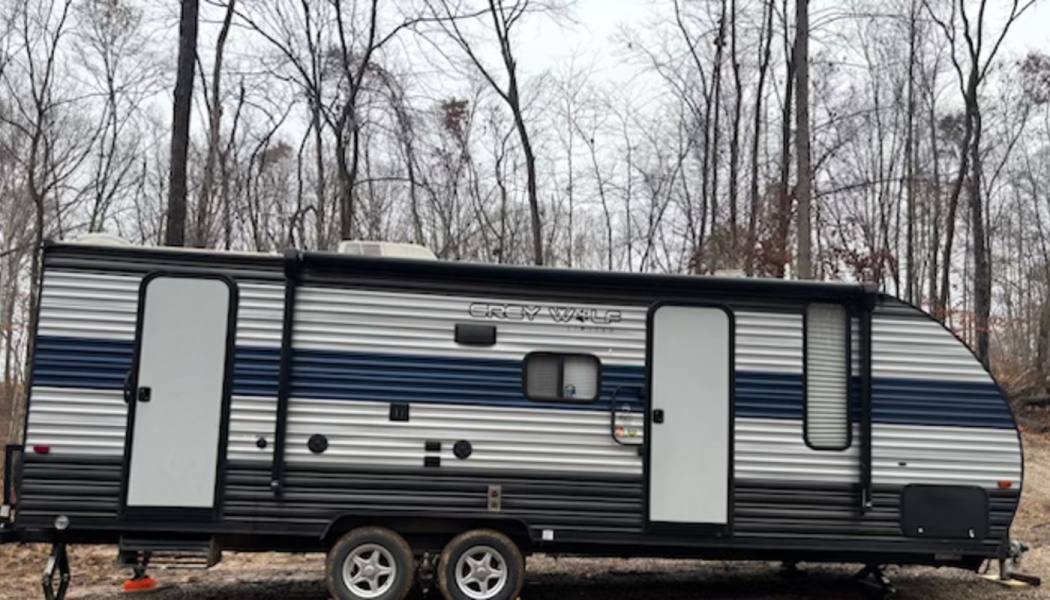 Grey Wolf travel trailer with silver siding and a blue stripe, parked in front of a wooded forest.