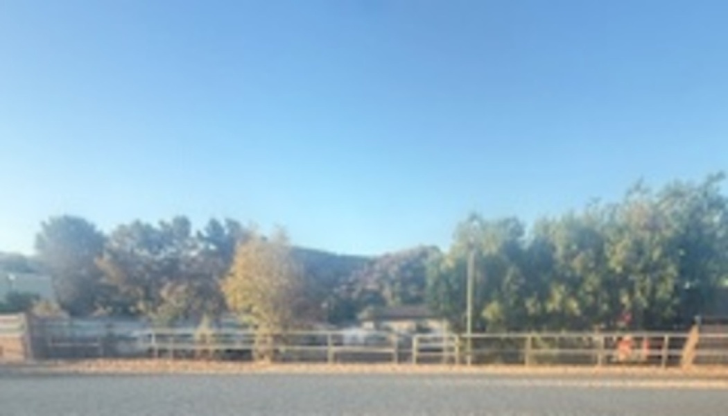 An outdoor riding arena with a wooden fence, surrounded by trees and hills under a clear blue sky.