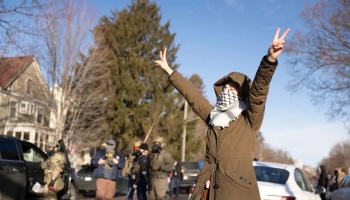 A person in a brown parka and face scarf holds up peace signs outdoors with people in tactical gear behind them.