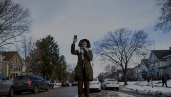 A woman in a hooded coat holds up her phone and shouts excitedly on a snowy residential street.