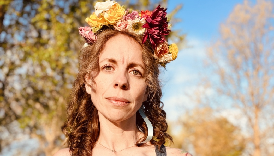 Woman with curly brown hair wearing a vibrant flower crown outdoors in warm sunlight.