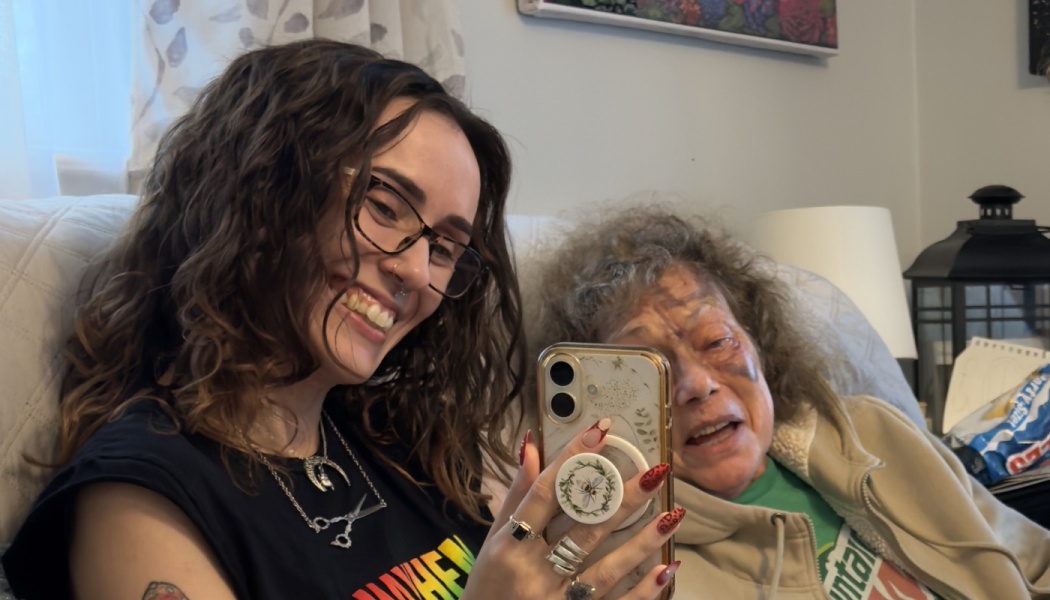 A young woman smiles while taking a smartphone photo with an elderly woman who has facial bruises.