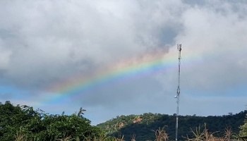 A vibrant rainbow arcs across a cloudy sky beside a tall telecommunications tower and green hills.