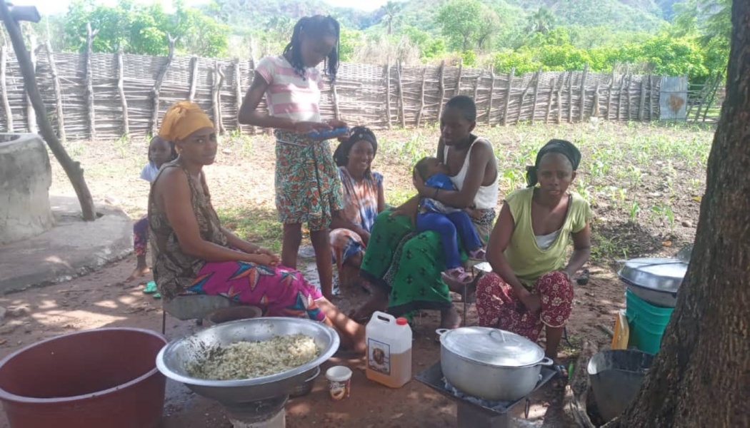 A group of women and children gather outdoors in a rural village to prepare food in large cooking pots.