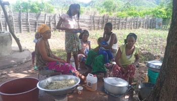 A group of women and children gather outdoors in a rural village to prepare food in large cooking pots.