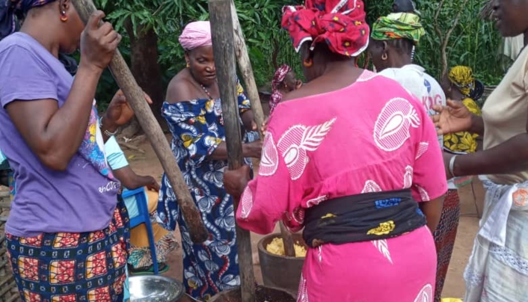 Women in vibrant, patterned dresses work together outdoors, using large wooden pestles to pound food in mortars.