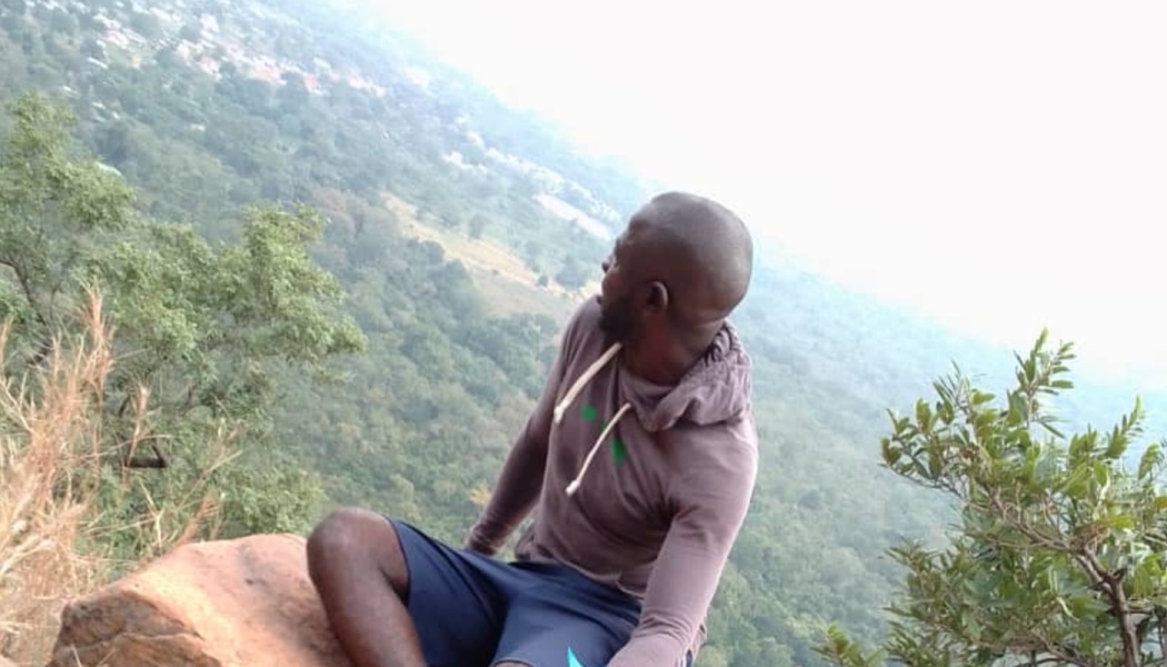 A man in a hoodie sits on a rocky cliff, looking out over a sprawling green valley.