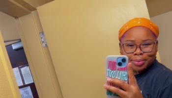 A smiling person wearing glasses and an orange headwrap taking a mirror selfie.