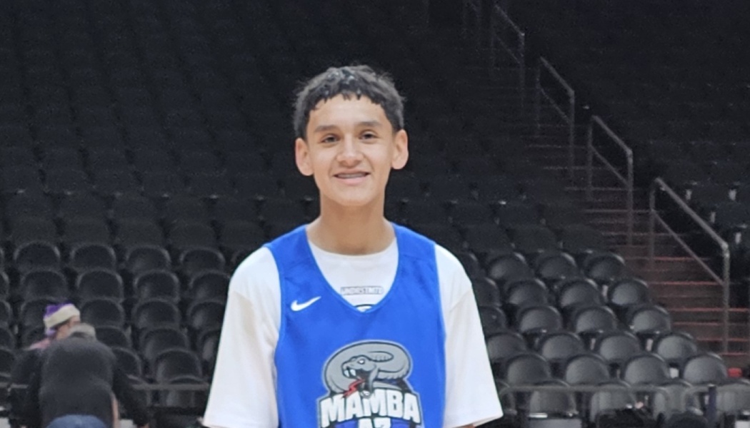 A smiling young basketball player in a blue Mamba jersey standing in front of empty arena seats.
