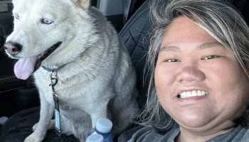 A smiling woman takes a selfie inside a car with a light-colored husky sitting in the seat next to her.