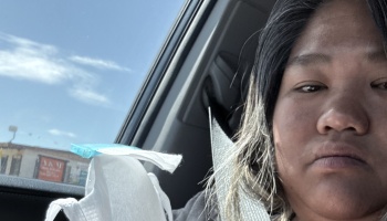 Close-up selfie of a person inside a car with a seatbelt on, looking tired with a white face mask nearby.
