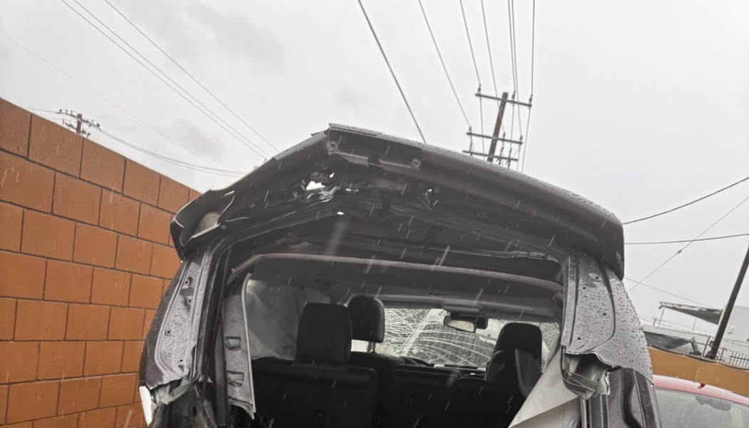Severely crushed roof and frame of a dark vehicle in falling rain with deployed airbags visible inside.