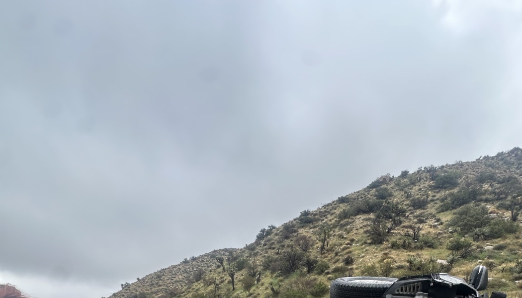 A damaged vehicle lies overturned on a scrubby desert hillside under a cloudy, grey sky.