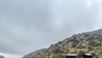 A damaged vehicle lies overturned on a scrubby desert hillside under a cloudy, grey sky.