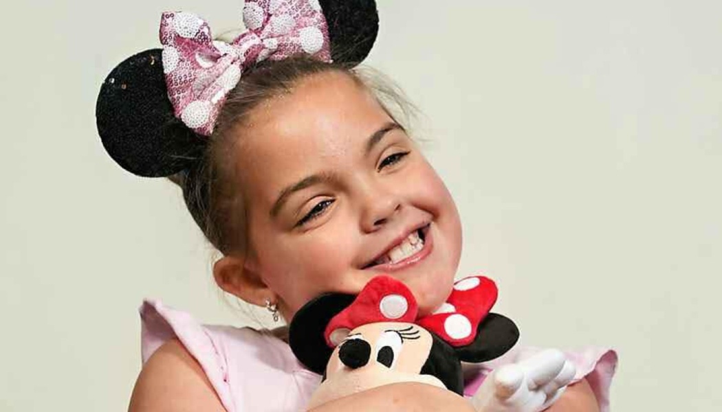 Smiling young girl wearing sparkly Minnie Mouse ears headband while hugging a Minnie Mouse plush toy.