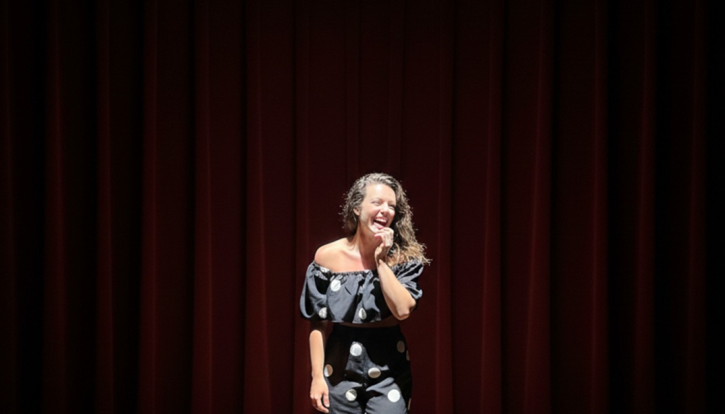 A smiling woman in a polka dot outfit stands under a spotlight in front of red theater curtains.