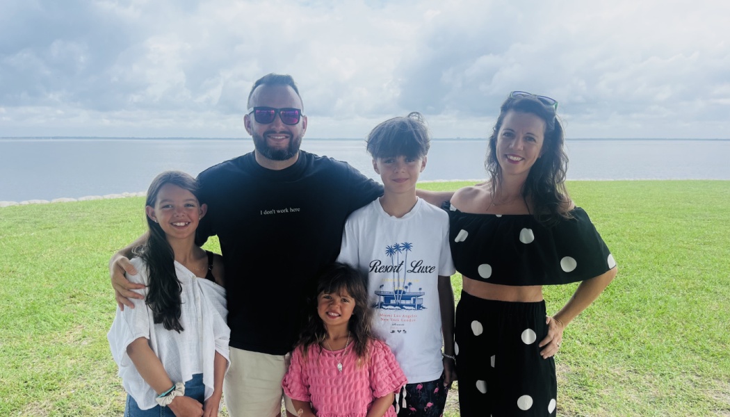 A smiling family of five standing together on a grassy lawn near water under an overcast sky.