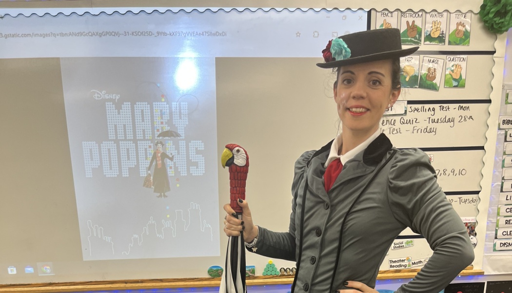 A teacher dressed as Mary Poppins holds a parrot umbrella next to a screen displaying Mary Poppins.
