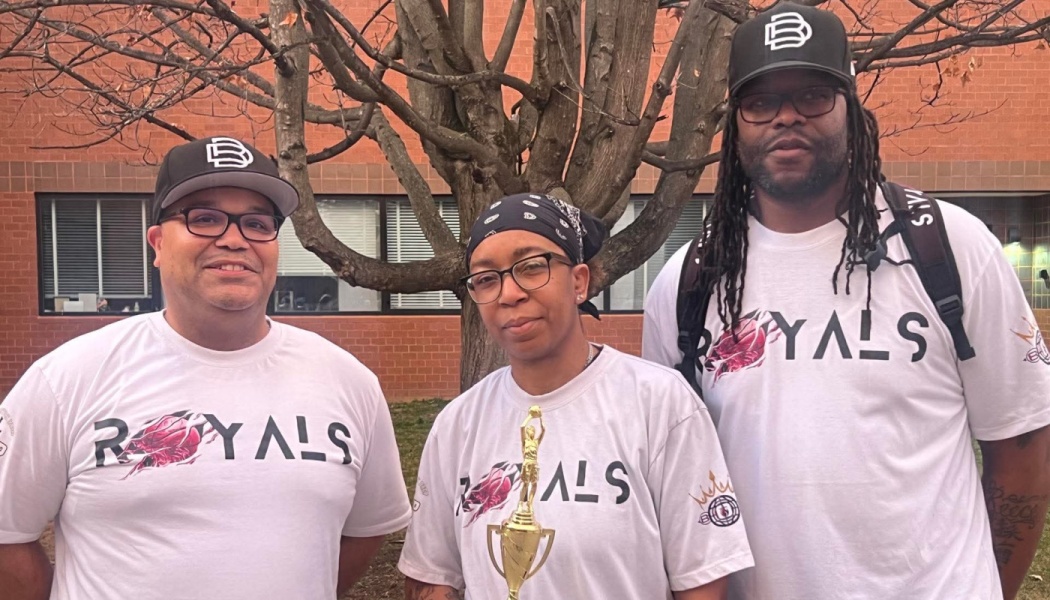Three people in matching white Royals t-shirts pose outside, with the woman in the middle holding a gold trophy.