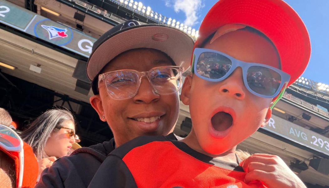 A smiling adult and an excited young boy in blue sunglasses posing together at a baseball stadium.