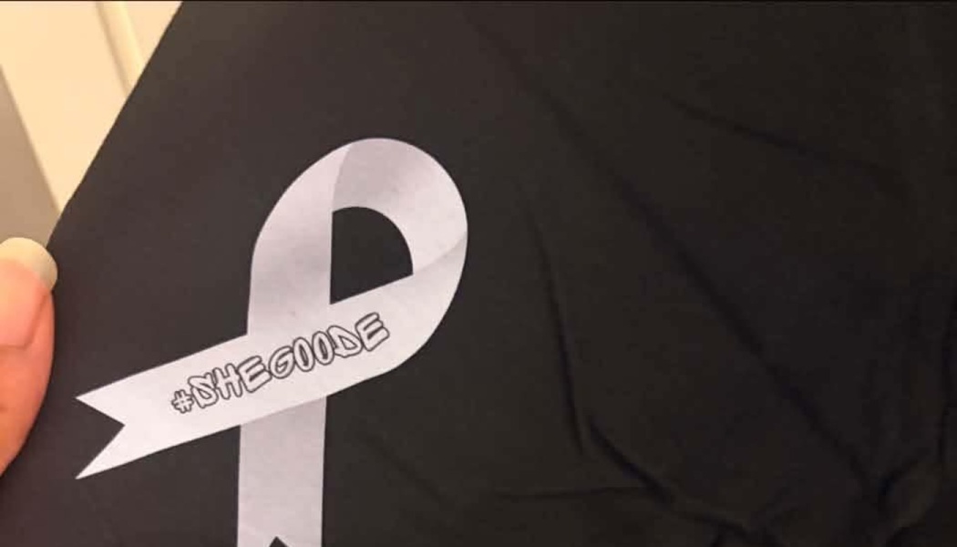 Light grey awareness ribbon printed on a black shirt, featuring the hashtag #SHEGOODE.