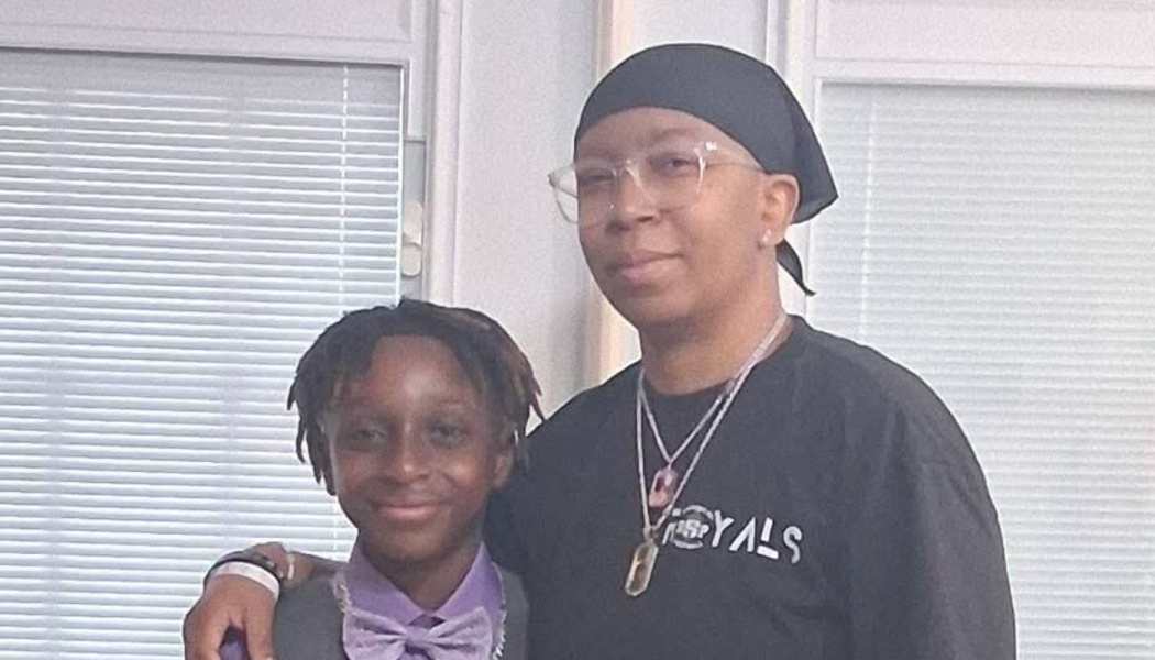 A smiling young boy in a bowtie standing next to an adult wearing glasses and a black headwrap.