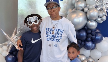 A person in an "Everyone Watches Women's Sports" t-shirt poses with two kids in front of blue and silver balloons.