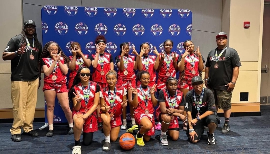A youth girls basketball team and their coaches pose with medals in front of a blue tournament backdrop.