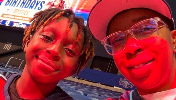 A child with dreadlocks and an adult in glasses smile under red arena lighting at a basketball game.