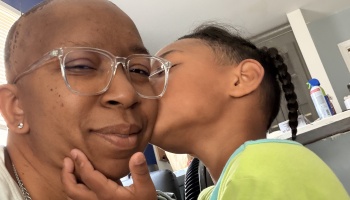 A young child affectionately kisses the cheek of a smiling Black adult wearing clear glasses.