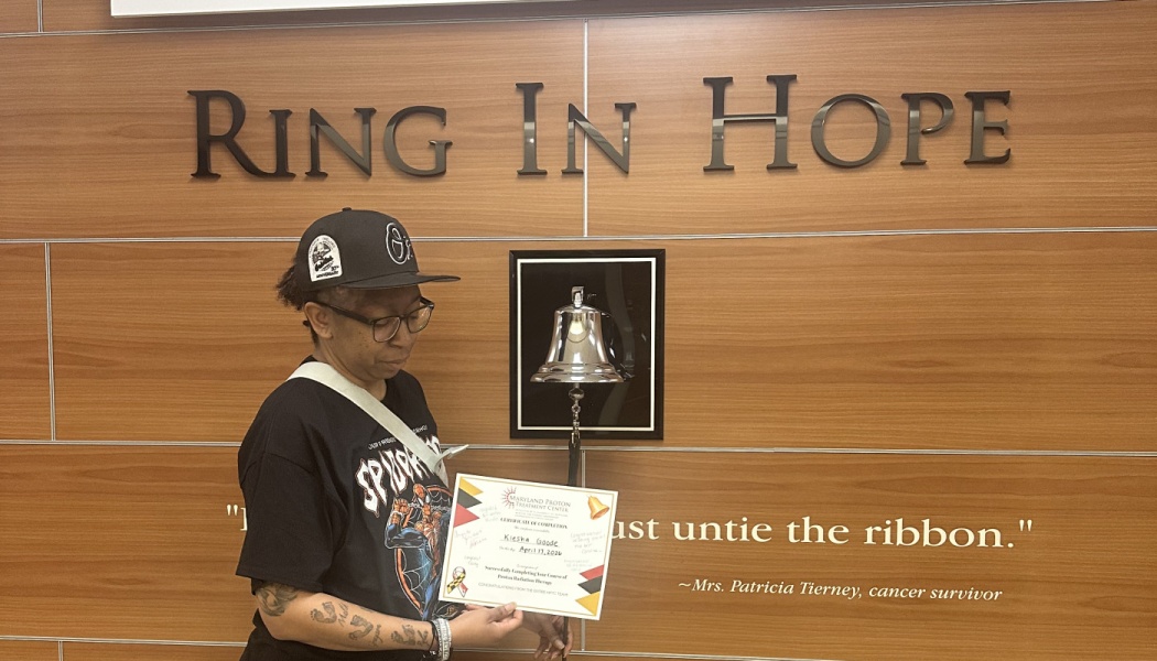 A woman holds a radiation therapy completion certificate in front of a "Ring In Hope" bell.