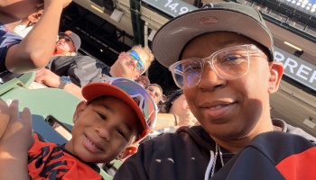 A smiling adult wearing glasses and a young child in a cap take a selfie together in the stands at a baseball game.