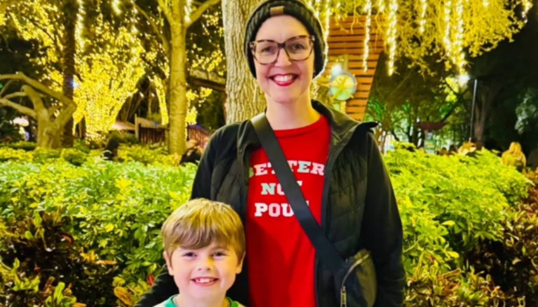A smiling woman in a beanie and glasses poses with a young boy in front of bright holiday lights at night.