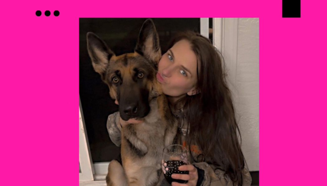 A woman with long dark hair poses beside a German Shepherd while holding a glass of red wine.