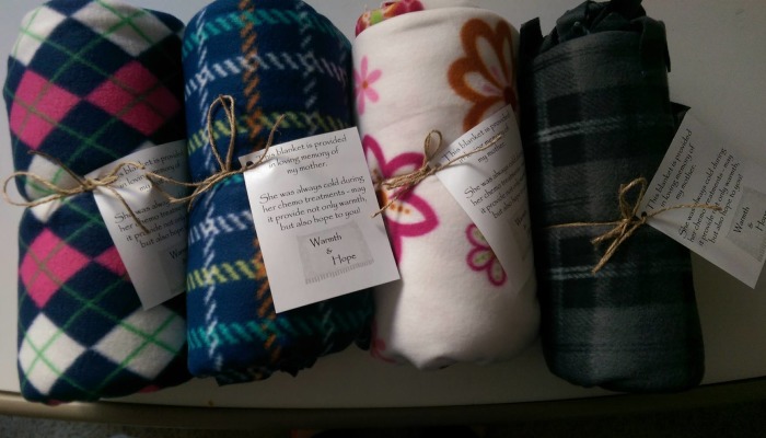 Four rolled patterned fleece blankets tied with twine, each with a memorial tag reading Warmth & Hope.