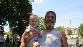 A smiling man with glasses and a goatee holding a cheerful baby boy outdoors on a sunny day.