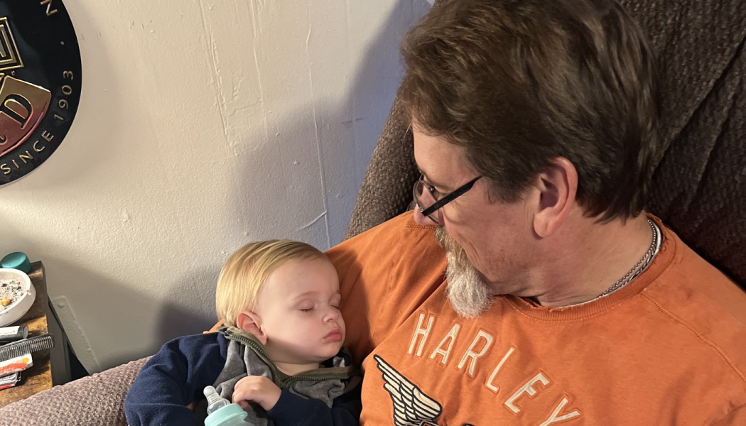 A man with glasses looks down lovingly at a sleeping blond toddler cradled in his arms.