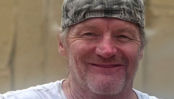 Close-up portrait of a smiling middle-aged man wearing a camouflage cap and white t-shirt.