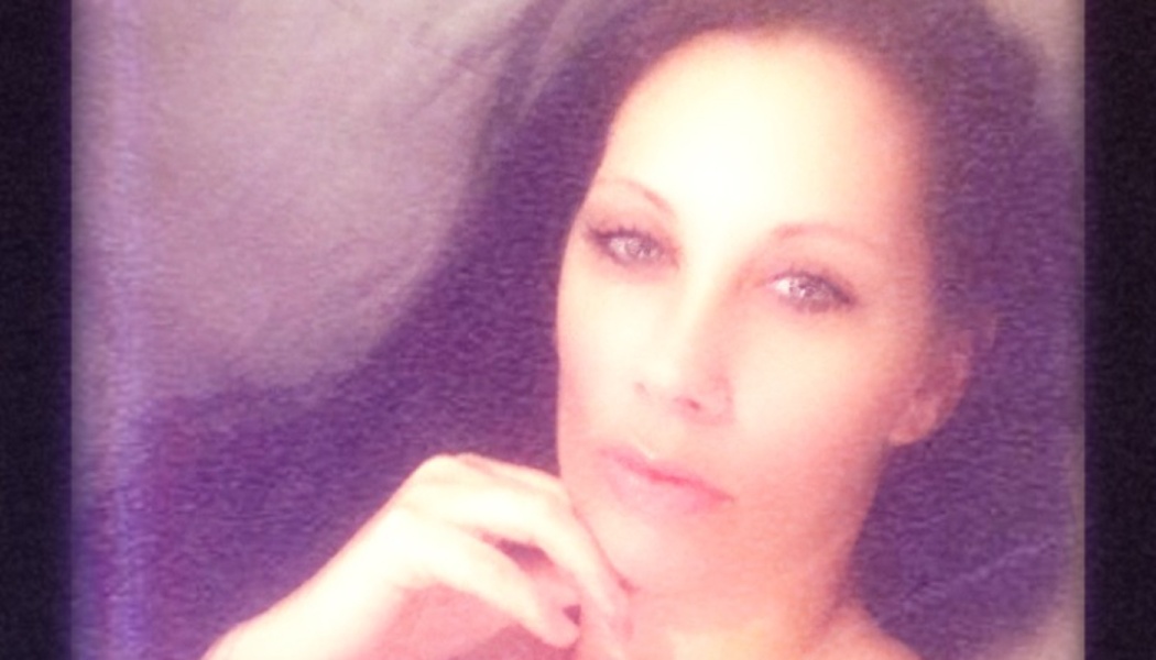 A woman with dark hair and light eyes resting her hand near her chin under soft purple lighting.