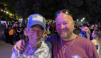 An older man in a blue hat and glasses smiles alongside a younger man with a goatee at an outdoor night event.