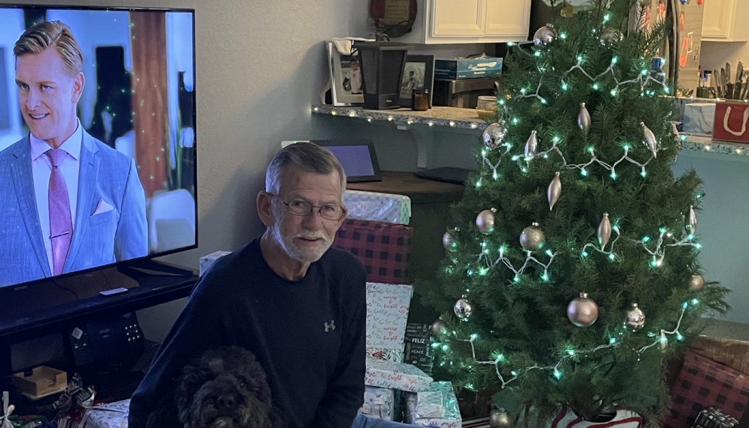 An older man with glasses and a black dog sit next to a decorated Christmas tree with wrapped presents.