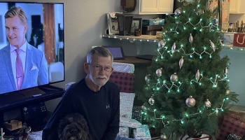 An older man with glasses and a black dog sit next to a decorated Christmas tree with wrapped presents.