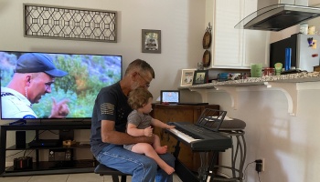 An older man holds a toddler on his lap while the baby plays an electric keyboard in a living room.