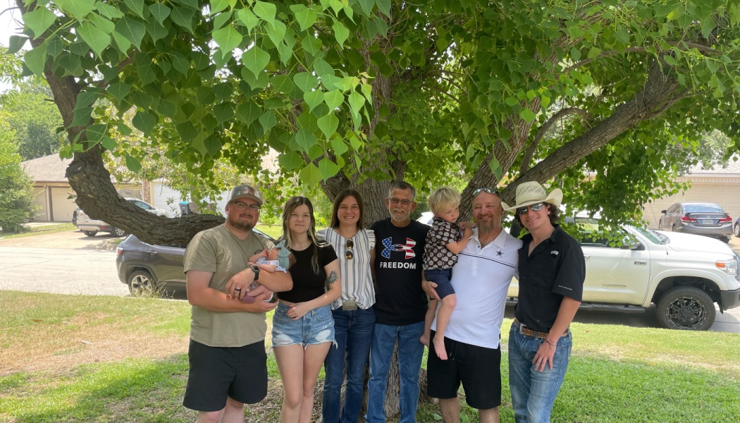 A smiling multi-generational family, including a newborn and toddler, standing together under a large leafy tree.