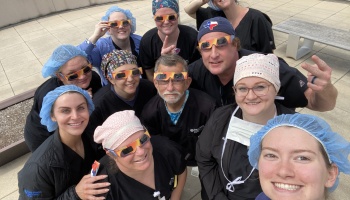 A smiling group of medical staff in scrubs wear solar eclipse glasses outdoors for a group selfie.