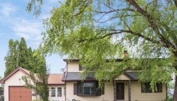 A two-story beige house with brown trim, an attached garage, and large green trees in the front yard.
