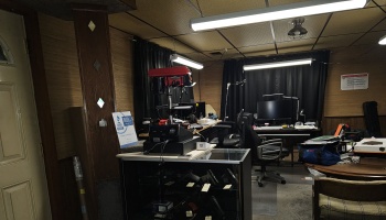 A workshop room with a red drill press on a glass display case, a computer desk setup, and wood-paneled walls.