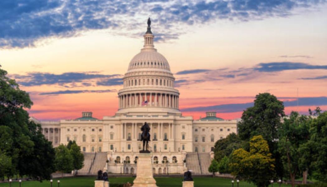 The US Capitol building framed by green trees against a colorful sunset sky with pink and blue clouds.